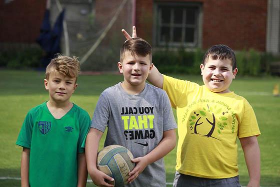 Three boys pose on a field with a soccer ball, smiling for the camera. One boy holds his fingers up as bunny ears to another.